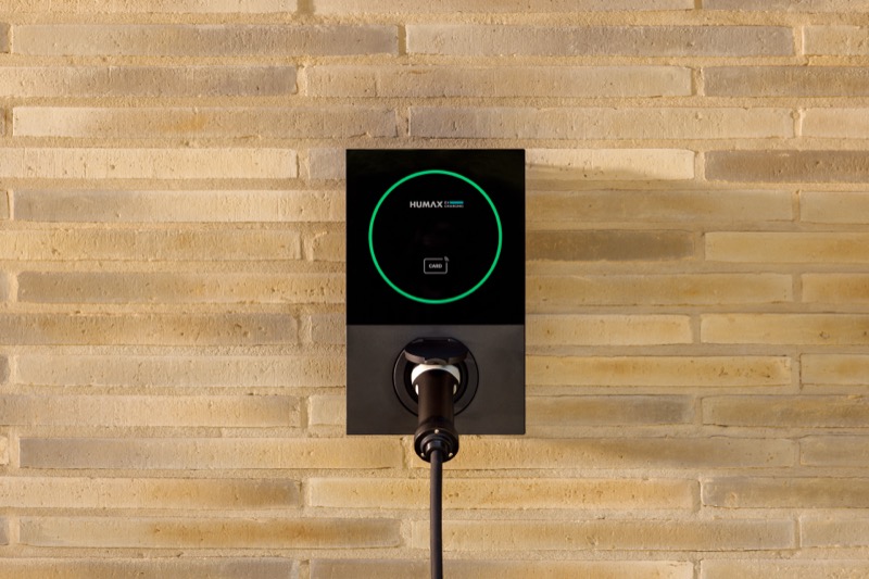 The Humax MX7 charger mounted on a stone-clad wall, glowing green to indicate ready to charge